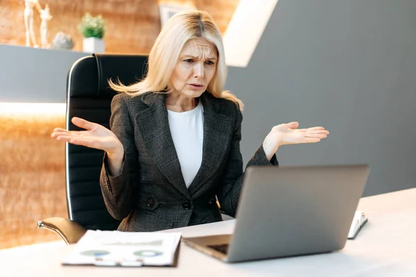 Discouraged upset middle aged blonde business woman, manager or broker, female executive, sit at the table in office, uses laptop, shocked by news or message, suffered financial loss,dissatisfied face