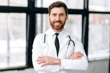 Kafkasyalı yakışıklı erkek doktor, terapist, cerrah, kardiyolog tıbbi üniformalı ve steteskoplu, hastanede duruyor, kameraya bakıyor, kollarını kavuşturup gülümsüyor.