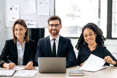 Başarılı çok ırklı iş adamları, modern ofiste oturan zarif yaratıcı meslektaşlar, proje üzerinde birlikte çalışıyorlar, dizüstü bilgisayar kullanıyorlar, finansal belgeleri analiz ediyorlar, kameraya bakıyorlar, gülümsüyorlar.