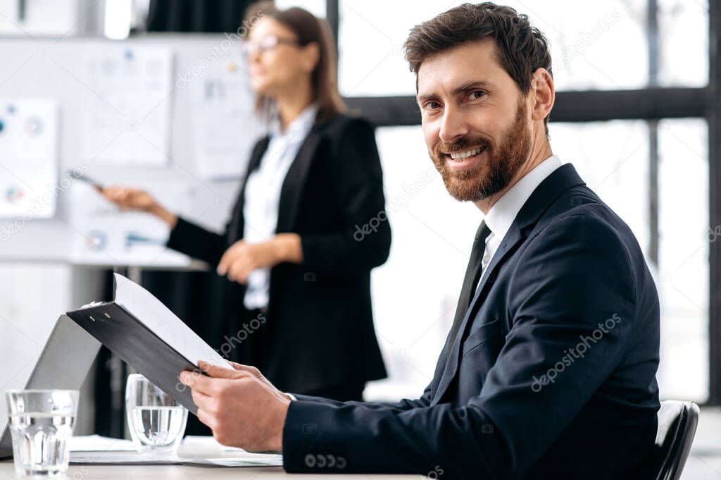 Successful business man, company ceo, sits in the office during corporate brainstorm, looks at camera, smiles. Portrait of attractive caucasian male business leader in formal suit at briefing meeting