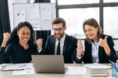 Zafer, zafer. Birlikte çalışan eğlenceli yaratıcı iş ortakları. Çok ırklı meslektaşlar birlikte strateji üzerinde kafa patlatıyorlar, modern ofiste oturuyorlar, dizüstü bilgisayar kullanıyorlar, iyi bir anlaşma yapıyorlar, kar ediyorlar, gülümsüyorlar.