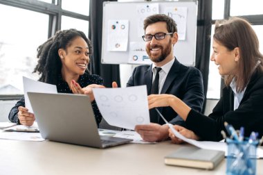 Başarılı yaratıcı iş ortakları bir proje üzerinde çalışıyor. Çok ırklı meslektaşlar birlikte strateji üzerine beyin fırtınası yapıyorlar, modern ofiste oturup, belgeler üzerinde çalışıyorlar, dizüstü bilgisayar kullanıyorlar, tartışıyorlar, gülümsüyorlar.