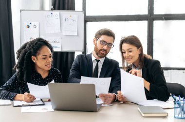 Bir projede iş ortakları birlikte çalışıyor. Çok ırklı meslektaşlar birlikte strateji üzerine beyin fırtınası yapıyorlar, modern ofiste oturup, belgeler üzerinde çalışıyorlar, dizüstü bilgisayar kullanıyorlar, tartışıyorlar, gülümsüyorlar.