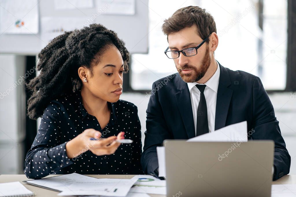 Business partners working together on a project. African american girl and caucasian man are brainstorming on strategy together, sitting in modern office, studying documents, using laptop, discussing