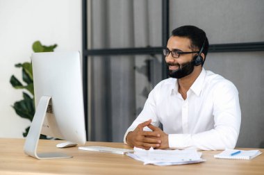 Zarif, çekici, zeki Hintli girişimci, iş adamı, satış müdürü, tüccar, kulaklıkla modern ofiste masada oturuyor, iş arkadaşlarıyla veya müşterileriyle video aracılığıyla konuşuyor, arkadaşça gülümsüyor.