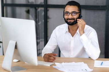 Gözlük ve kulaklık takan yakışıklı, pozitif zekalı Hintli bir adamın portresi, yardım hattı uzmanı, çağrı merkezi operatörü, doğrudan kameraya bakıyor, dostça gülümsüyor. Hizmet desteği kavramı
