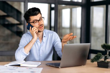 Modern bir ofiste dizüstü bilgisayarın arkasında otururken, akıllı telefondan konuşan, resmi tişörtlü başarılı bir adam telefonla konuşuyor, eliyle işaret veriyor.