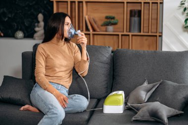 Kafkasyalı esmer kadın astım spreyiyle hasta. Sağlıksız bir kadın evde solunum yapıyor. Tedavi için nebulizör ve sprey kullanıyor. Evdeki kanepede oturuyor.