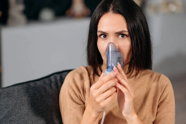 Close-up foto van zieke blanke jonge vrouw met een inhalator. Ongezond brunette meisje doet inhalatie thuis, ze gebruikt vernevelaar en inhalator voor de behandeling, zitten op de bank, kijkt naar de camera — Stockfoto