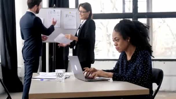 Joven mujer de negocios afroamericana bonita y positiva sentada en el lugar de trabajo en la oficina moderna, durante la lluvia de ideas de negocios con colegas, mirando a la cámara, sonriendo amigable — Vídeos de Stock