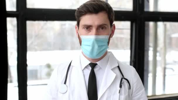 Orgulloso guapo médico caucásico de medicina general, de pie en el hospital, quitándose la máscara médica de la cara, mirando a la cámara, sonriendo. Fin de la cuarentena y restricciones — Vídeo de stock