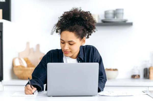 Distance learning, online lesson. Focused smart african american female student, in stylish casual wear, studying remotely, using a laptop, taking notes during online lecture, sitting at home