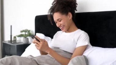 Gadget ve teknoloji konsepti. Mutlu Afro-Amerikan genç bayan akıllı telefonunu evde yatarken kullanıyor, cep telefonuyla mesajlaşıyor, internetten haberleri okuyor, sosyal medyada geziniyor, gülümsüyor.