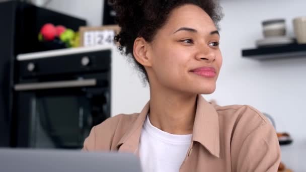 Jolie jeune femme afro-américaine, pigiste, spécialiste de l'informatique, assise à la maison dans la cuisine, regardant ailleurs, rêvant, continue d'utiliser un ordinateur portable pour le travail, envoyant des SMS à ses collègues, travaillant sur un projet, souriant — Video