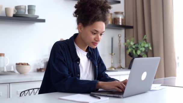 Cute successful joyful young african american woman, freelancer, IT specialist, sitting at home in the kitchen, using laptop for work, texting with colleagues, working on a project, smiling happily — ストック動画