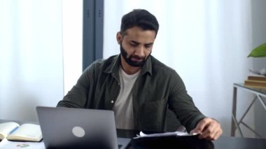 Konsantre olmuş zeki Hintli ya da Arap işadamı, tüccar, simsar, modern bir ofiste bir iş yerinde oturuyor, istatistiklere odaklanıyor, finansal tablolara bakıyor, dizüstü bilgisayarda çalışıyor, notlar alıyor.