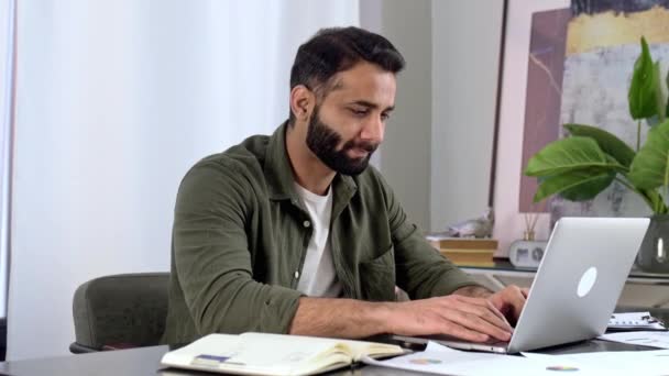 Homme intelligent et positif indien ou arabe en tenue décontractée, pigiste ou manager, assis au bureau au bureau, écoutant un webinaire, prenant des notes, apprenant, améliorant ses compétences, souriant — Video