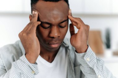 Close-up of exhausted african american guy in stress, sits at home alone, in casual clothes, experiencing fatigue, headache, migraine, massaged temples, closes eyes, needs rest, medicines, treatment
