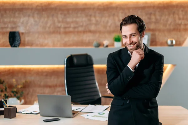 Portrait of an elegant caucasian successful businessman in formal stylish suit, entrepreneur, company ceo, standing in his modern office near workdesk, looking at the camera, smiling joyfully — Foto de Stock