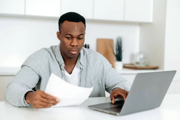 Concentrated african american man, freelancer, stock trader, designer, sitting at the table at home, in casual wear, looks into a laptop, analyzes online reporting, profit, working on a project — 스톡 사진