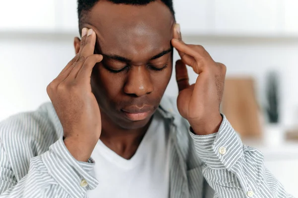 Close-up do afro-americano exausto em estresse, senta-se em casa sozinho, em roupas casuais, experimentando fadiga, dor de cabeça, enxaqueca, templos massageados, fecha os olhos, precisa de descanso, medicamentos, tratamento — Fotografia de Stock