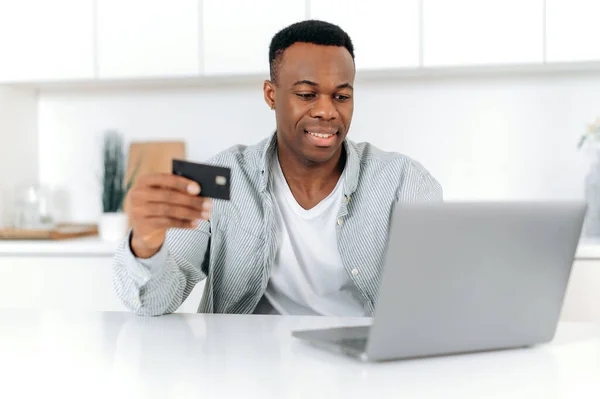 Happy african american man sitting at home, dressed in casual wear, using laptop and credit card for online shopping, entering data for payment, online transaction, order home delivery, smiling — Foto de Stock