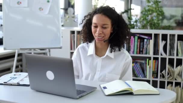 Avlägsen support, affärsmöte. Afroamerikansk positiv ung kvinna med headset, sitta på modernt kontor, prata med kollegor eller ha online samråd med kunder via videosamtal — Stockvideo