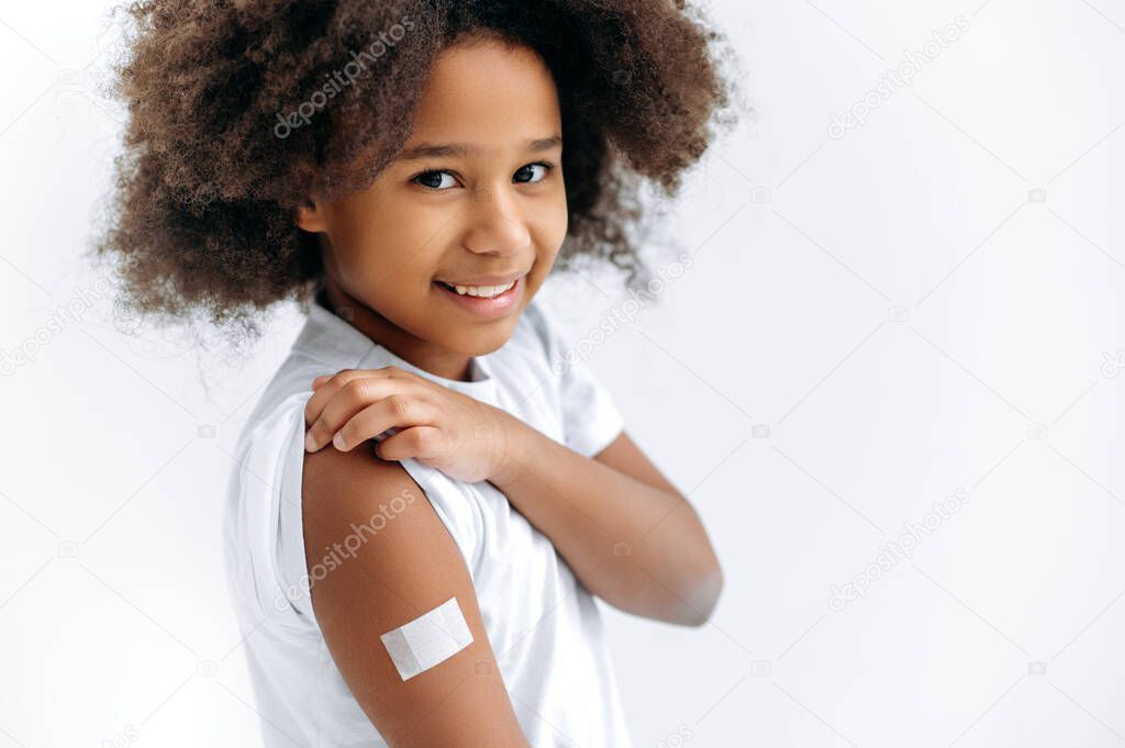 Close-up of African American cute little girl, preschooler, with band-aid on shoulder, received a vaccine, protection against covid19, and other diseases, stands on an isolated white background, smile