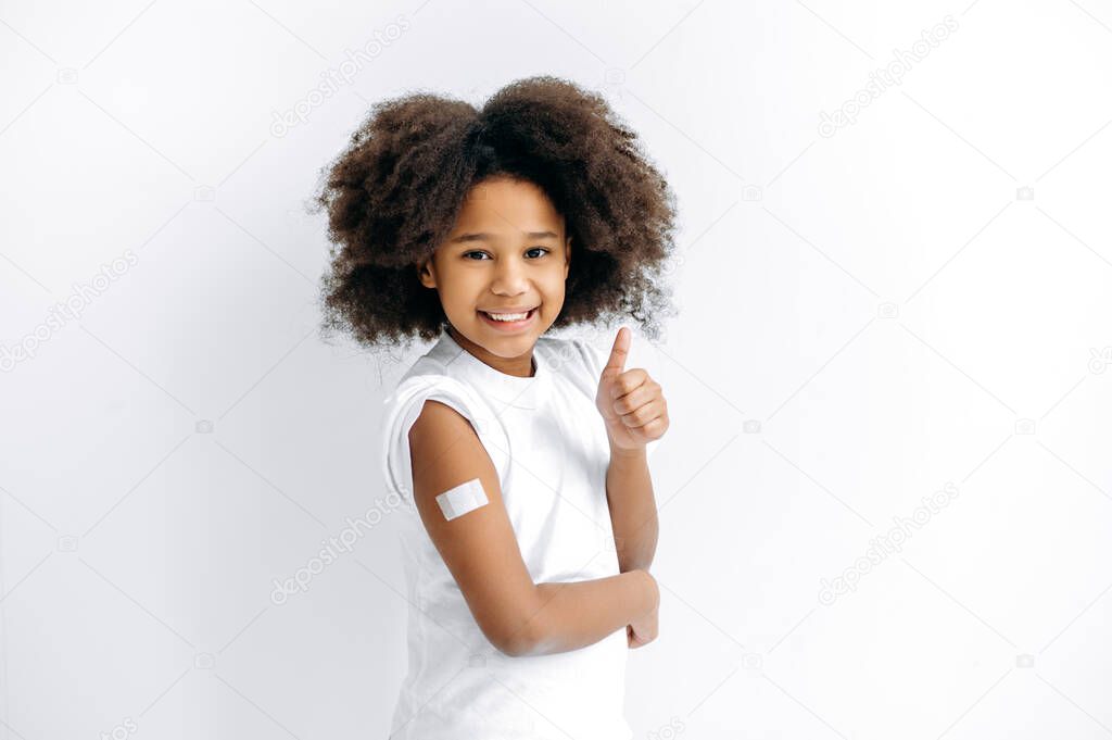 Positive African American little girl, preschooler, with band-aid on shoulder after vaccination, for immunity against covid19, and other diseases, stand on isolated white background, thumb-up gesture