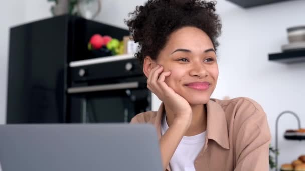 Beautiful African American curly haired girl, designer, student, freelancer, uses a laptop at home in the kitchen, works on the Internet, looks away, thinks, dreams, smiles happily, working on project — ストック動画