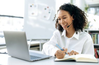 Kulaklığı, menajeri, destek çalışanı, online danışmanı olan arkadaş canlısı Afrikalı Amerikalı kız. Ofiste dizüstü bilgisayarda oturuyor, notlar alıyor, müşterilerle çevrimiçi görüşmeler yapıyor, gülümsüyor.