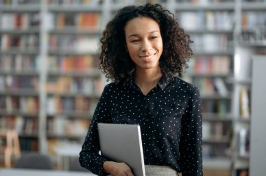 Kendinden emin, Afro-Amerikan kıvırcık saçlı kadın, bilişim uzmanı, tüccar, yönetici, modern bir ofiste şık iş elbiseleriyle duruyor, elinde bir dizüstü bilgisayar tutuyor, yüzünü çeviriyor, gülümsüyor.