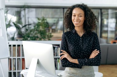 Güzel başarılı afro-amerikan kıvırcık iş kadını, pazarlamacı, avukat modern ofiste masanın yanında duruyor, şık iş kıyafetleri içinde, kollarını kavuşturmuş, kameraya bakıyor, dostça gülümsüyor.
