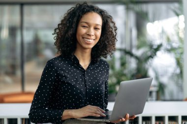 Portre başarılı, esmer kıvırcık saçlı kız, finans analisti, denetçi, bilişim uzmanı, ofiste duruyor, iş kıyafetleri içinde, elinde açık bir laptop tutuyor, kameraya bakıyor, gülümsüyor.