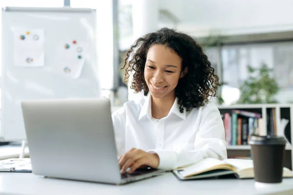 Charmant Afro-Amerikaans meisje, met krullend haar, financieel adviseur, makelaar, auditor, zit achter een laptop, chat met collega 's of medewerkers, kijkt naar het scherm, glimlacht — Stockfoto