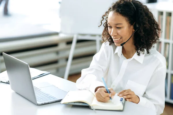 Vänlig positiv afroamerikansk kvinna med lockigt hår, online försäljningschef, support servicearbetare, sitter vid ett skrivbord framför en bärbar dator i moderna kontor, gör arbetsanteckningar — Stockfoto