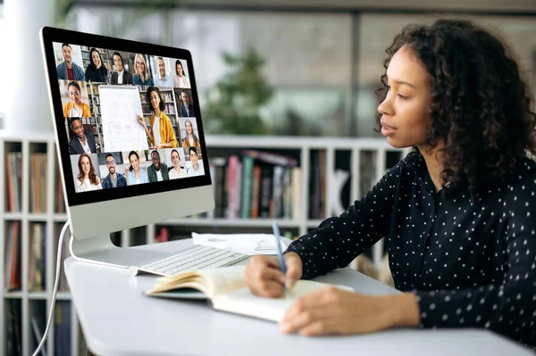 Онлайн обучение. Умная африканская кудрявая американка, сидит за рабочим столом перед экраном компьютера, концентрируется на часах и слушает онлайн лекцию, делает заметки, получает знания — стоковое фото
