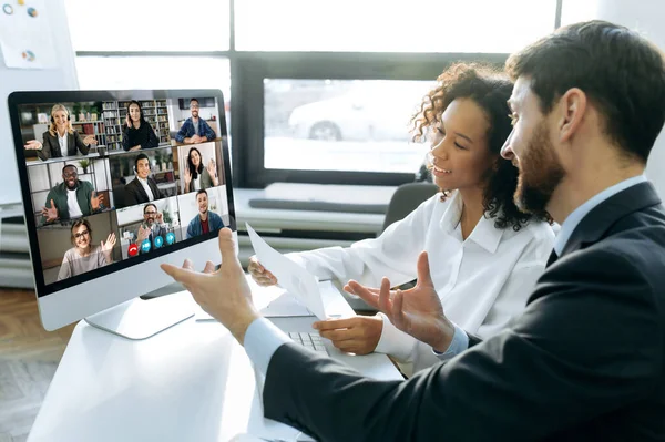 Business colleagues, co-founders of the company, sit at a table in the modern office in front of a computer screen, hold an online video conference with international colleagues, brainstorming