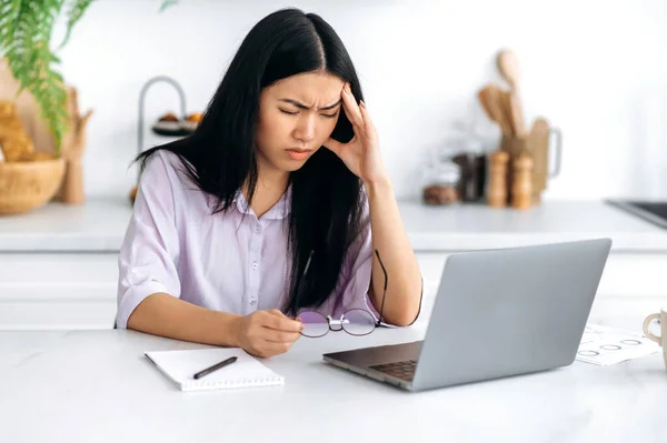 Unavená z práce na notebooku smutná asijská mladá žena, na volné noze nebo obchodník s akciemi, sedí u laptopu v kuchyni, frustrovaná vyčerpaná dívka drží hlavu, bolí ji hlava, migréna, zavírá oči — Stock fotografie