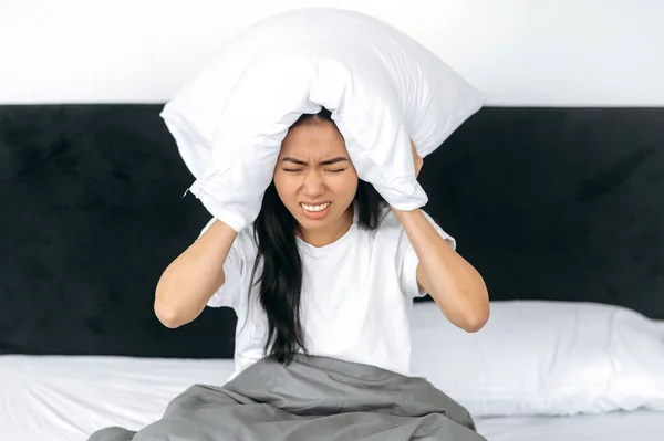 Geïrriteerde Aziatische meisje, zitten in een bed thuis, bedekt haar oren met een kussen, probeert te slapen, sluit zich af van lawaai, heeft een slechte droom, rusteloze droom. Slaap en rust concept — Stockfoto