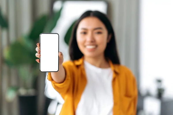 Gadis Cina yang tidak fokus, mengenakan pakaian santai, berdiri di rumah di ruang tamu, memegang smartphone di tangannya dengan layar mockup putih kosong untuk iklan, melihat ke kamera, tersenyum — Stok Foto