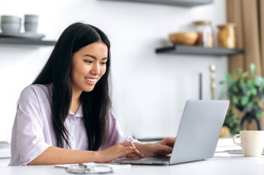 Şık giyinen güzel, zeki Çinli kız, serbest çalışan, öğrenci ya da yaratıcı tasarımcı, mutfakta oturuyor, dizüstü bilgisayar kullanıyor, bir proje üzerinde çalışıyor, internette geziniyor, gülümsüyor.