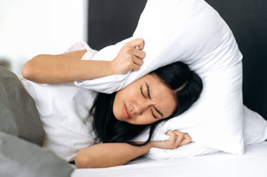 Uyku ve dinlenme konsepti. Üzgün Çinli kız, evde bir yatakta uzanıyor, kulaklarını yastıkla örtüyor, uyumaya çalışıyor, kendini gürültüden koruyor, kötü bir rüya görüyor, huzursuz bir rüya.