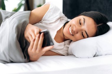 Mutlu Asyalı genç esmer kadın akıllı telefonunu evde yatakta yatarken kullanıyor, akıllı telefonla mesajlaşıyor, internetten haberleri okuyor, sosyal medyada geziniyor, mutlu bir şekilde gülümsüyor.