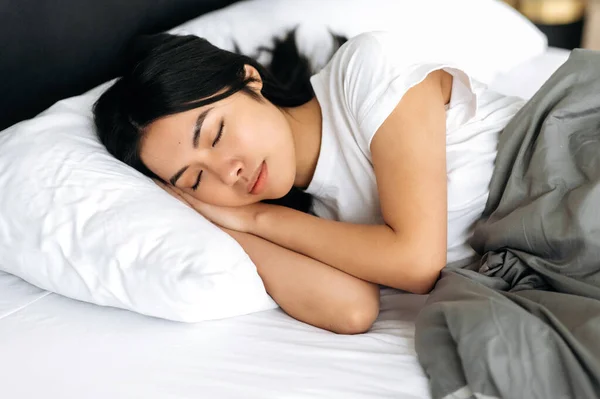 Zijaanzicht van een slapend Aziatisch meisje in een gezellig bed in appartement, aangename dromen, gezonde slaap. Mooie Chinese vrouw in wit t-shirt onder de deken in bed in de ochtend, ziet zoete dromen — Stockfoto