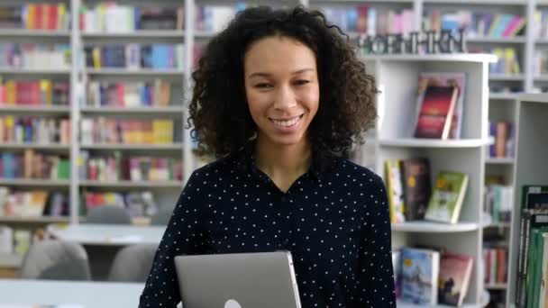 Positiva chica afroamericana encantadora con el pelo rizado, vestida con una camisa, estudiante o profesor, sosteniendo un ordenador portátil, caminando por la biblioteca, mirando a la cámara, sonriendo amigable — Vídeos de Stock