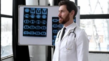 Gururlu beyaz erkek tıp doktoru, cerrah, pulmonolog, tıbbi üniformalı, omuzlarında gözlük ve steteskop ile klinikte durup kameraya bakıp gülümsüyor.