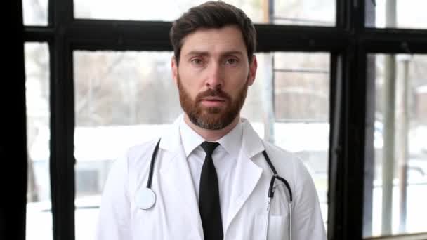 Θλιβερός κουρασμένος εργατικός άνδρας γιατρός γενικής ιατρικής, με λευκή ιατρική στολή με στηθοσκόπιο στους ώμους του, στέκεται σε ένα νοσοκομείο κοντά στο παράθυρο, κοιτάζοντας την κάμερα, φαίνεται εξαντλημένος — Αρχείο Βίντεο