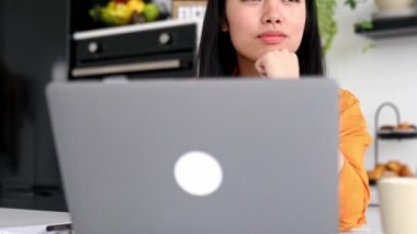 Güzel, pozitif Asyalı esmer kadın, tasarımcı, serbest çalışan ya da öğrenci, mutfakta dizüstü bilgisayarla oturuyor, çalışıyor ya da uzaktan çalışıyor, yandan bakıyor, düşünüyor, hayal ediyor, gülümsüyor.