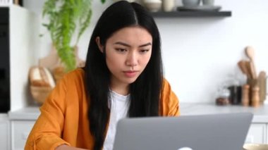 Başarılı, mutlu duygusal Çinli kız, şık kıyafetler içinde, serbest çalışan, bilişim uzmanı, mutfakta dizüstü bilgisayarla oturmuş, büyük kazanç, zafer, iyi haber, yumruklarla el kol hareketi, gülümseme...